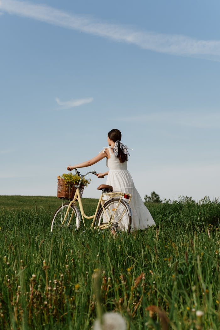 A woman in a white dress stands in a meadow with a vintage bicycle filled with flowers.